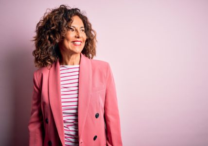 Middle age beautiful businesswoman wearing elegant jacket over isolated pink background looking away to side with smile on face, natural expression. Laughing confident.